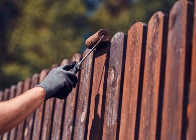 Man in protective gloves is painting wooden fence in bright summer day.